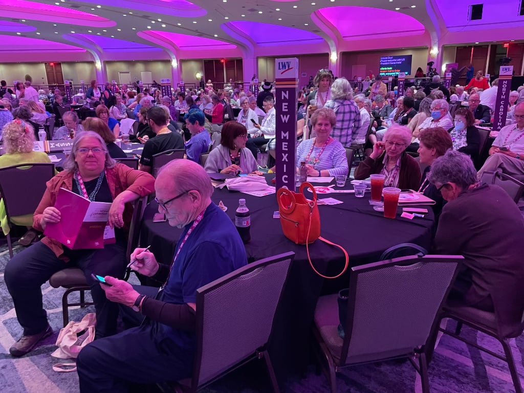 A group of conference attendees sit inside a large conference room. A vertical sign reading "New Mexico" is visible in the center.